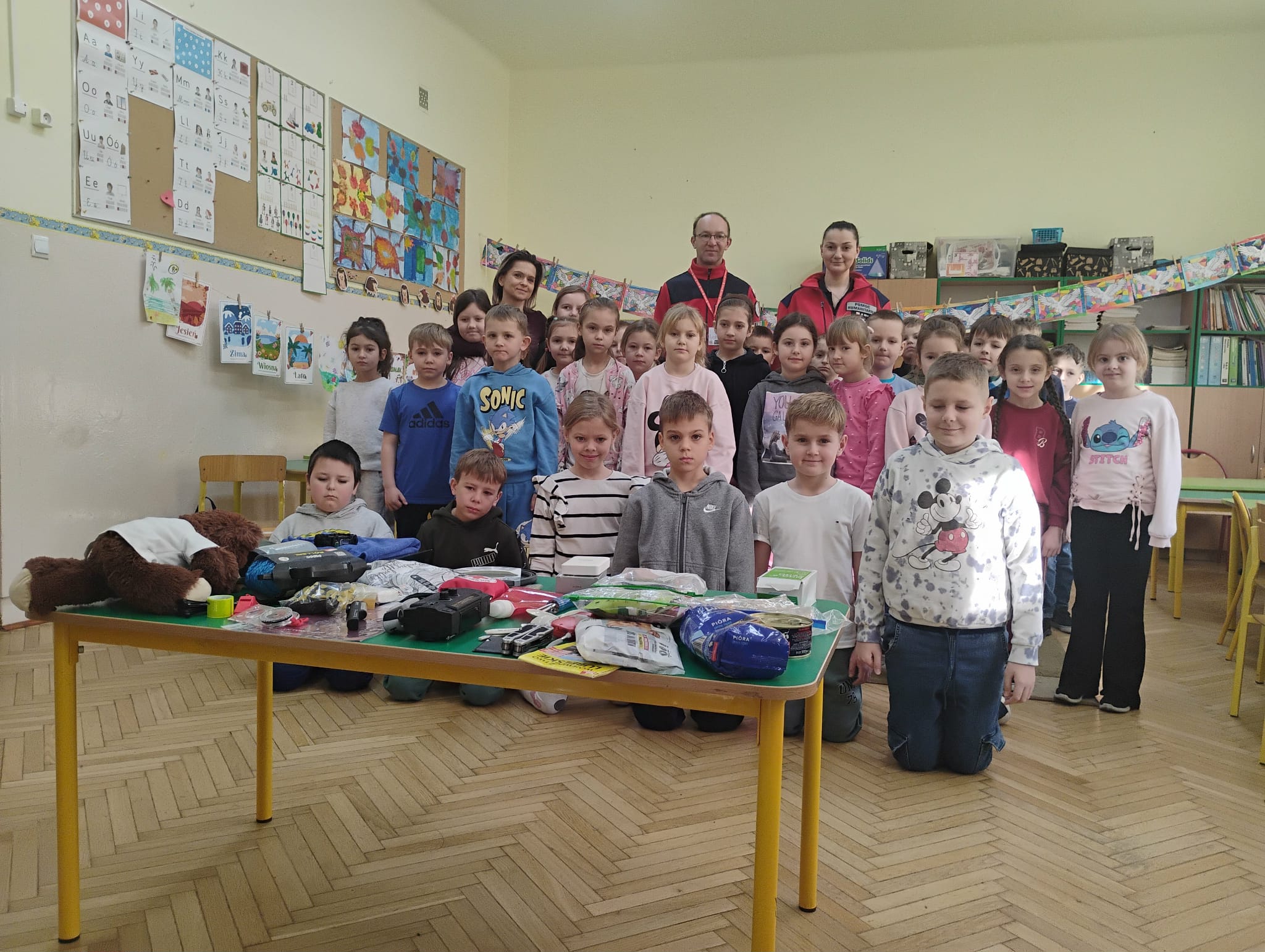Na tym zdjęciu uczniowie pozują do wsp&oacute;lnego zdjęcia wraz z dwojgiem ratownik&oacute;w w czerwonych uniformach. Cała grupa stoi za stołem, na kt&oacute;rym rozłożone są liczne elementy plecaka ewakuacyjnego: środki higieniczne, opatrunki, latarki, woda, żywność oraz inne praktyczne akcesoria. Dzieci stoją w dw&oacute;ch rzędach, a w tle widać dekoracje i prace plastyczne wiszące na ścianach sali lekcyjnej.